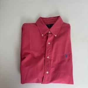 Rosa skjorta från Ralph Lauren XL - Snygg rosa skjorta från Ralph Lauren i bomull med klassisk krage och knappar framtill. Långärmad modell med blå broderad logga på bröstet. Perfekt för dig som gillar stilrena och färgstarka plagg.