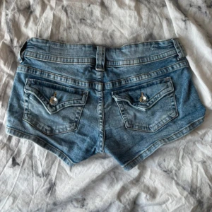 Blå jeansshorts från Bershka - Snygga ljusblå jeansshorts från Bershka med dubbla knappar fram och klassiska fickor både fram och bak. Bakfickorna har lock med silvriga knappar. Shortsen har låg midja och är gjorda i mjukt denimtyg, perfekta för varma dagar.