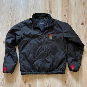 Svart vindjacka Peak Performance S - Svart vindjacka från Peak Performance med broderad logga på bröstet i rött, orange och gult. Jackan har dragkedja framtill, elastisk midja och röda detaljer med knapp vid ärmsluten. Tillverkad i polyester och fodrad med polyamid och Thermolite.