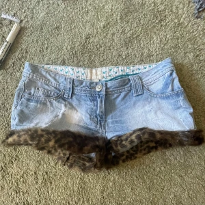 Lågmidjade jeansshorts med leopardpäls - Unika ljusblå jeansshorts från Miso med låg midja och leopardmönstrad pälsdetalj längs bensluten, silverfärgade nitar vid fickan och knappstängning framtill. Säljer för att de är förstora för mig </3