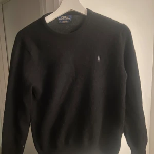 Svart junior merinoulltröja Ralph Lauren - Junior Ralph Lauren i mjuk merinoull. Rund hals och diskret broderad logga på bröstet. Perfekt för dig som gillar stilrena och tidlösa plagg med premiumkänsla. Bred: 38 Längd: 58