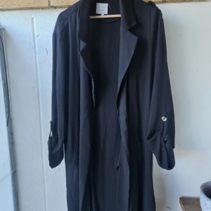 Svart lång kappa från Club XPRS - Stilren svart lång kappa från Club XPRS med klassisk krage och knappar på ärmarna. Jackan har en rak passform och är perfekt för dig som gillar en clean och enkel look. Passar till många olika outfits och har snygga detaljer vid ärmsluten.