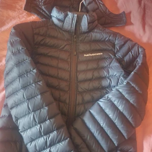 Svart dunjacka Peak Performance - Snygg svart dunjacka från Peak Performance med quiltad design och hög krage. Jackan har dragkedja framtill och diskret logga på bröstet. Perfekt för kyliga dagar och enkel att matcha med allt.