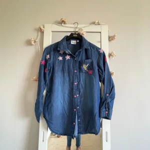 Disney Tink jeansskjorta - Snygg jeansskjorta från Disney Tink i blå denim med broderade rosa och lila stjärnor samt Tinkerbell-detalj på bröstfickan. Skjortan har långa ärmar, rosa knappar och en relaxed passform. Perfekt för dig som gillar unika detaljer och magisk stil.storlek står för 13-14 år ,men  man kan säga dom en vanlig smal storlek 