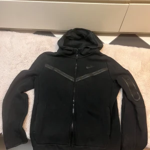 Svart Nike hoodie med dragkedja  - Svart hoodie från Nike i storlek XL barnstorlek, med hel dragkedja framtill och huva. Snygg detalj med svarta ränder över bröstet och en praktisk ficka med dragkedja på ena ärmen. Perfekt för chill eller träning. säljer för den är för stor