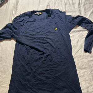 Mörkblå långärmad tröja Lyle & Scott - Mörkblå långärmad tröja från Lyle & Scott med klassisk gul logga på bröstet. Tröjan är i mjuk bomull och har en enkel, stilren design som passar perfekt till jeans eller chinos.