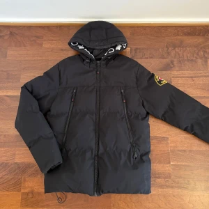 Svart pufferjacka från Zavetti Canada - Svart pufferjacka från Zavetti Canada med huva och feta logodetaljer på insidan. Jackan har två snedställda dragkedjefickor framtill, patch på ärmen och justerbar dragsko nedtill. Perfekt för kalla dagar och riktigt snygg streetstil.