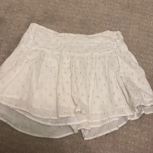 Vit kortkjol med guldiga detaljer Gina Tricot - Supersöt vit kortkjol från Gina Tricot med små guldiga broderade detaljer. Kjolen har volang och resår i midjan för en bekväm passform. Perfekt för sommarens alla tillfällen och lätt att matcha med olika toppar.