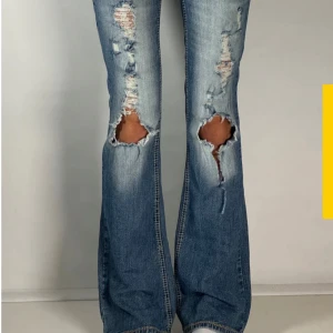 Blå slitna bootcut jeans med broderade fickor, ultra low waist. - Säljer ett par blå bootcut jeans med slitningar och stora hål vid knäna. Jeansen har snygga broderade bakfickor och låg midja. De är tillverkade i klassiskt denimtyg och har en cool, avslappnad vibe.