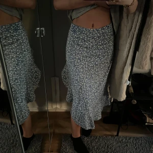 Blommig blå omlottkjol med volang - Säljer en blå långkjol med vitt blommigt mönster. Kjolen har omlott-design med volang längs nederkanten och en dragkedja bak för enkel påklädning. Materialet känns lätt och flowy, perfekt för dig som gillar en feminin och trendig look.