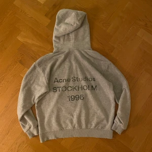 Acne Studios hoodie - Grå hoodie från Acne Studios med tryck på ryggen: 'Acne Studios STOCKHOLM 1996'. Klassisk modell med huva, ribbade muddar och avslappnad passform. Tillverkad i 100% bomull för en mjuk och skön känsla. Perfekt för en chill och stilren look. Strl M. Osäker på äkthet därav priset, passar på tjejer och killar