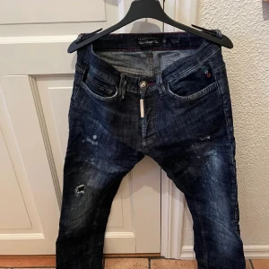 Mörkblå jeans från Philipp Plein - Snygga mörkblå jeans från Philipp Plein, med slitningar och coola detaljer som ger en exklusiv och trendig look. Jeansen har klassisk femficksdesign, silverfärgad knapp med logga, dragkedja samt tydliga kontrastsömmar som förstärker designen.  Inköpta direkt i Philipp Pleins butik och endast använda ett par gånger. Äkthetsintyg samt originalbox medföljer.