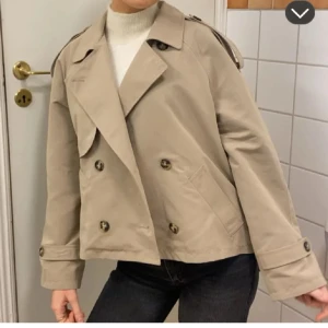 Beige trench coat - Snygg beige trench coat med dubbelknäppning och bred krage. Jackan är kortare i modellen och har dekorativa axelklaffar samt knappar vid ärmslut. Perfekt för dig som gillar en stilren och trendig look.