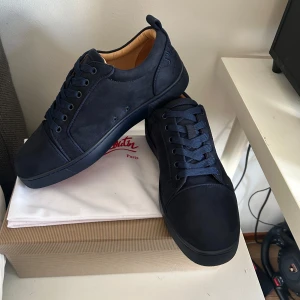 Mörkblå sneakers från Christian Louboutin - Snygga mörkblå sneakers från Christian Louboutin i mocka med klassisk röd sula. Skorna har rund tå, platt sula och ton-i-ton snörning. Insidan är ljusbeige och baksidan har broderad logga. Perfekta för dig som vill sticka ut med exklusiva detaljer.