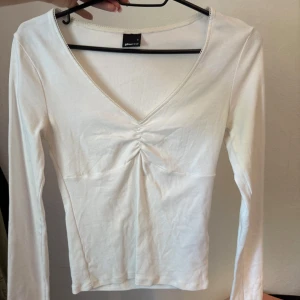 Vit långärmad topp från Gina Tricot - Snygg vit långärmad topp från Gina Tricot med v-ringning och rynkdetalj framtill. Toppen är i mjuk bomull och har en figurnära passform som passar perfekt till jeans eller kjol.
