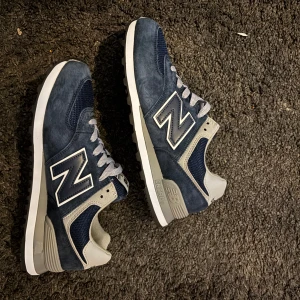New Balance 574 Classic blå/grå sneakers - Säljer ett par New Balance 574 Classic sneakers i marinblått och grått. Skorna har meshpaneler, mockadetaljer och stora N-loggor på sidorna. Grå snörning och vit/grå sula ger en clean look. Perfekta för dig som gillar retrostil och streetwear.