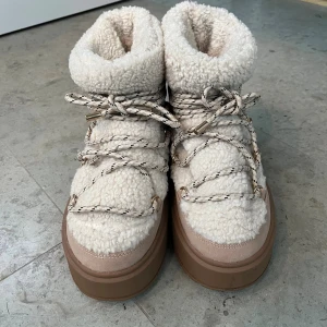 Beiga moon boots från Bershka - Supermysiga moon boots från Bershka i beige teddy-material med breda snören och grov platåsula. Skorna har detaljer i mocka och syntet, samt coola guldfärgade metalldetaljer på snörningen. Perfekta för kalla dagar när du vill vara både varm och trendig.