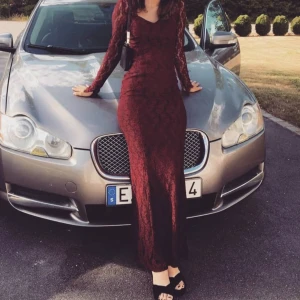 Vinröd balklänning - Vinröd långklänning i figurnära modell med vacker spets över hela klänningen. Klänningen har långa ärmar, v-ringad urringning och dragkedja i ryggen. Perfekt för bal eller festliga tillfällen. Materialet är spets med fodrad insida för en bekväm känsla.