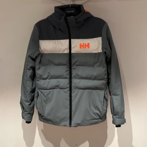 Grå pufferjacka Helly Hansen 176/16 - Snygg pufferjacka från Helly Hansen i olika grå blå nyanser med vit panel och orange HH-logga. Jackan har huva, hel dragkedja och justerbara ärmslut. Perfekt för kalla dagar och har en sportig vibe med tydlig branding på både fram- och baksida. Väldigt bra som skidjacka.
