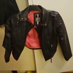 Svart skinnjacka Vintage Style 158 - Cool svart skinnjacka från Vintage Style med quiltade detaljer på axlarna, dragkedja framtill och snygga tryckknappar. Jackan är croppad och har ett rosa innerfoder som ger extra edge. Perfekt för dig som vill ha en rockig look.