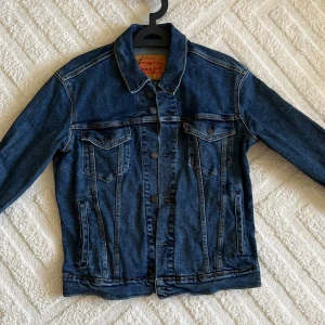 Levi's klassisk blå jeansjacka S - Säljer en klassisk blå jeansjacka från Levi's i storlek S. Jackan har snygga knappar framtill, två bröstfickor med lock och sidofickor. Materialet är 98% bomull och 2% elastan för lite stretch. Perfekt med krage och coola sömmar.