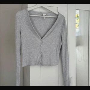 Grå ribbad kofta från H&M - Snygg grå ribbad kofta från H&M i storlek M. Koftan har v-ringad hals, långa ärmar och knappar framtill. Croppad passform som ger en trendig look, perfekt att styla med jeans eller kjol. Mjuk och skön kvalitet.
