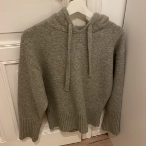 Grå stickad hoodie från Zara - Mysig grå hoodie från Zara i stickat material. Tröjan har huva med snörning, långa ärmar och breda ribbade muddar vid ärmslut och nederkant. Perfekt för kyliga dagar och enkel att matcha med jeans eller leggings.