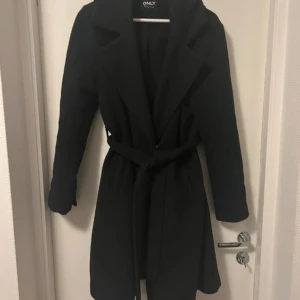 Svart kappa från ONLY med bälte - Stilren svart kappa från ONLY med klassisk krage och knytskärp. Tjockare, perfekt för vintern! Storlek M men skulle säga att den passar S också! Nypris runt 600-700
