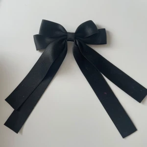 Svart rosett diadem i satin - Stilren svart rosett i satin med långa band, perfekt att använda som diadem eller håraccessoar. Ger en elegant och trendig look till din outfit. Rosetten har en klassisk form och en snygg glansig finish.