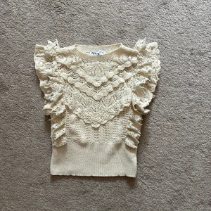 Beige stickad blus med volang från Zara - Supersöt beige stickad blus från Zara med volangdetaljer på axlar och bröst. Blusen har ett vackert spetsmönster och är ärmlös, perfekt för vår och sommar. Ger en romantisk och trendig look till din outfit!