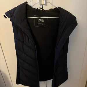 Svart dunväst med huva från Zara - Svart quiltad dunväst från Zara med dragkedja och stor, mysig huva. Västen har en stilren design med lätt figursydd passform och är perfekt för lager-på-lager. Passar dig som vill ha en enkel och snygg look under kyliga dagar.