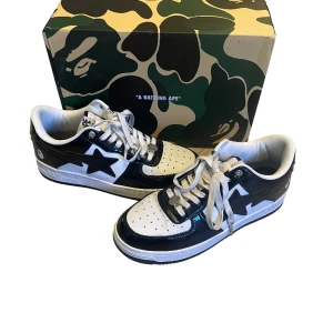 BAPE STA svartvita sneakers - Säljer ett par BAPE STA sneakers i svart och vitt med klassisk stjärndesign på sidan. Skorna har snörning, rund tå och platt sula. Tillverkade i glansigt syntetmaterial och levereras med originalkartong, dustbag och tag. Perfekta för dig som gillar streetwear och vill sticka ut.