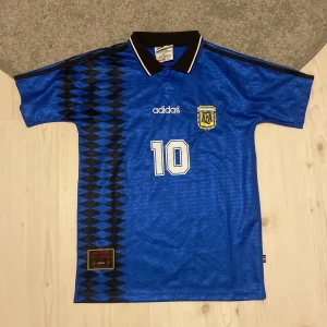 Argentina Maradona 10 blå Adidas tröja - Klassisk blå Argentina fotbollströja från Adidas med Maradona och nummer 10 på ryggen. Tröjan har svart krage, vita detaljer och ett unikt svart geometriskt mönster längs ena sidan. Broderad AFA-logga och Adidas-tryck framtill. Tillverkad i polyester.