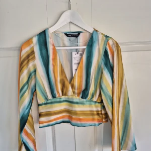 Blus/topp från Zara - Färgstark croppad blus från Zara i randigt mönster. Djup v-ringning och långa ärmar som är något vidare nedtill. Tillverkad i ett lätt, glansigt material som känns lyxigt och svalt. Helt oanvänd med lapparna kvar. Storlek S.