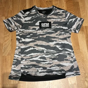Camouflage t-shirt från Björn Borg - Snygg t-shirt från Björn Borg med grått och beige camouflage-mönster framtill och svarta paneler på ryggen. Stor DPM-logga på bröstet. Perfekt för träning eller chill. Klassisk passform och korta ärmar.