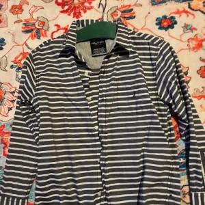 Randig skjorta från Nautica, marinblå - Snygg skjorta från Nautica i marinblått och vitt med horisontella ränder. Klassisk krage, bröstficka och knäppning framtill. Tillverkad i 100% bomull för en skön känsla. Perfekt för dig som gillar en stilren och avslappnad look.