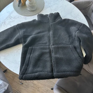 Svart teddyjacka Carin Wester S - Mysig svart teddyjacka från Juna Pile i storlek S. Jackan har hög krage, hel dragkedja framtill och två stora fickor. Tillverkad i fluffigt fleece-material som håller dig varm under kyliga dagar. Perfekt för dig som gillar en chill och avslappnad stil.