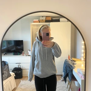 Grå hoodie med huva och ficka - En basic grå hoodie med stor huva och dragsnören. Tröjan har en klassisk känguruficka framtill och är i en loose passform. Perfekt för chill dagar och enkel att matcha med allt. Mjuk insida och ribbade muddar vid ärmslut och nederkant.