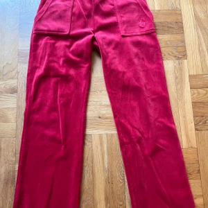 Röda Juicy Couture del ray i strl XS - Röda velourbyxor från Juicy Couture med raka ben, stora fickor fram och bak med knappar. Byxorna har en mjuk resår i midjan och klassisk Juicy Couture-logga på ena fickan. Perfekta för en chill och trendig look. Obs! Snöret i midjan är borta. Väldigt fint skick och endast använda några gånger.