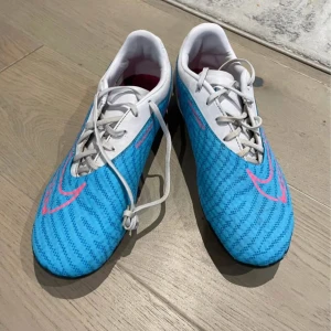 Nike Phantom fotbollsskor blå/rosa - Säljer ett par Nike Phantom fotbollsskor i blått och vitt med rosa detaljer och snörning. Skorna har en smal passform, platt sula med dobbar och är tillverkade i syntetmaterial. Perfekta för dig som vill sticka ut på planen med stilrena och sportiga skor.