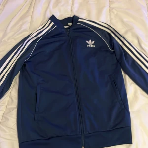 Blå Adidas track jacket med vita ränder - Säljer en klassisk blå Adidas track jacket med vita ränder längs ärmarna och Adidas-logga på bröstet. Jackan har hel dragkedja, ståkrage och två fickor framtill. Perfekt för en sportig och avslappnad stil.