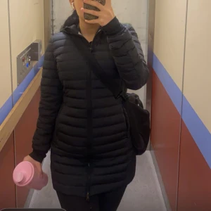 Parajumpers irene  - Säljer en svart lång dunjacka med huva och horisontella sömmar. Jackan har dragkedja hela vägen och en clean, stilren look som passar perfekt när det är kallt ute. Snygg passform och enkel att matcha med allt.