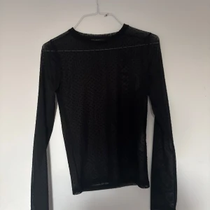 Svart mesh topp med prickigt mönster - Säljer en svart mesh topp med lång ärm och diskret prickigt mönster. Toppen är genomskinlig och har en tight passform med rund halsringning. Perfekt att styla lager på lager för en trendig look.