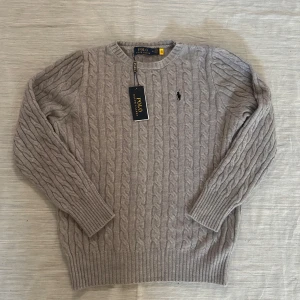 Grå kabelstickad tröja Polo Ralph Lauren - Kabelstickad tröja från Polo Ralph Lauren i en snygg ljusgrå nyans. Klassisk rund halsringning, ribbade muddar och ärmar. Tröjan har den ikoniska broderade loggan på bröstet och är tillverkad i mjuk ull för en lyxig känsla. Kvitto finns.