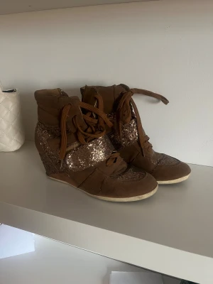 Bruna sneakers med glitter och kilklack - Säljer ett par bruna sneakers med kilklack och glittriga detaljer. Skorna har snörning och är tillverkade i mocka med inslag av syntetmaterial. Glittriga partier på sidorna och framtill ger dem en extra cool vibe. Perfekta för dig som vill sticka ut.