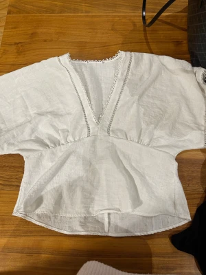 Vit blus i linne med spetsdetaljer - Säljer en vit blus i linne med v-ringad hals och fina spetsdetaljer längs kanten. Blusen har breda, korta ärmar och en lätt rynkad midja som ger en avslappnad siluett. Perfekt för dig som gillar en enkel men ändå söt stil.