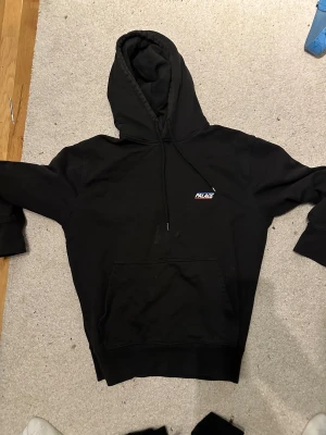 Palace hoodie - Svart hoodie från Palace med liten logotyp på bröstet. Klassisk design med huva och snörning, samt stor magficka framtill. Perfekt för dig som gillar streetwear och vill ha en enkel men snygg look.