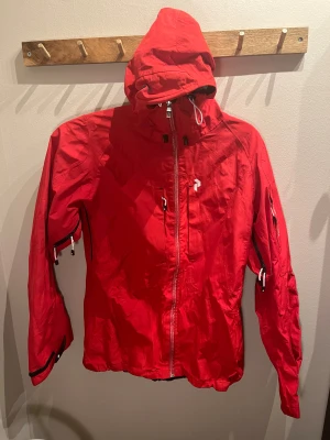 Peak Performance: röd skaljacka - Röd skaljacka från Peak Performance med huva,  dragkedja, flera praktiska fickor med dragkedjor och justerbara ärmslut. Det enda jag hittar är att dragkedjan fram är torkad (?) - därav pris. I övrigt bra skick. 