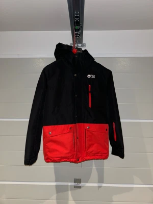 Svart och röd vinterjacka från Picture - Jacka från Picture i svart och rött med huva och flera fickor framtill. Jackan har dragkedja och tryckknappar, samt logga på bröstet. Perfekt för kalla dagar och har en sportig look med färgblockdesign. Med superbra fickor, med en duk för göra rent goggles. Storlek xs picture storlek 14=164cm 