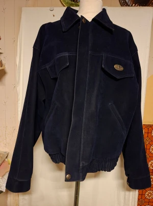 Mörkblå mockajacka från V Import Export - Snygg mörkblå mockajacka från V Import Export, made in Italy. Jackan har klassisk bombermodell med stora fickor framtill, bred krage och elastisk midja. Stängs med knappar och har detaljer i metall. Perfekt för dig som gillar retrovibbar och vill sticka ut.